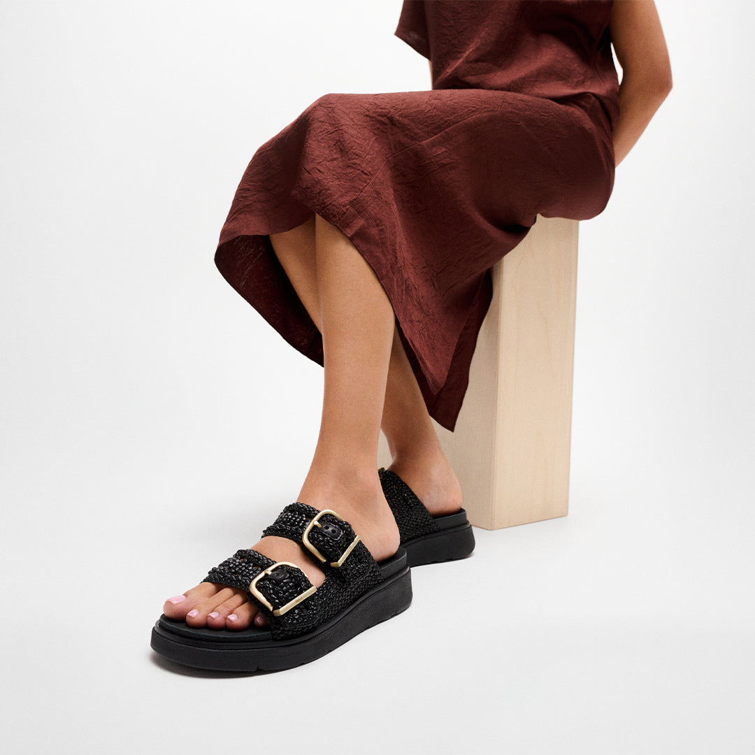Woman wearing FitFlop GEN-FF Woven Two-Bar Slides with woven straps, adjustable buckles and supportive cushioned sole.