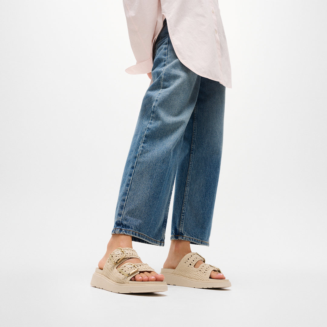 Woman wearing FitFlop GEN-FF Woven Two-Bar Slides with woven straps, adjustable buckles and supportive cushioned sole.
