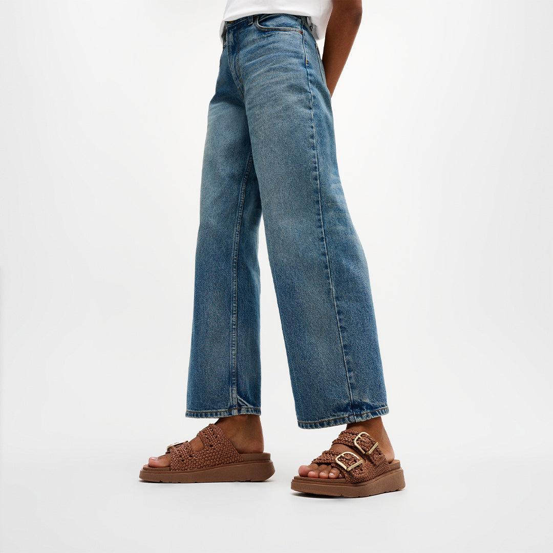 Woman wearing FitFlop GEN-FF Woven Two-Bar Slides with woven straps, adjustable buckles and supportive cushioned sole.